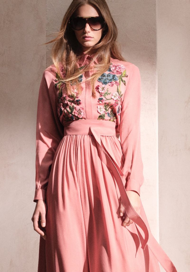 A fashionable adult female poses in a long-sleeve, ankle-length dress with floral embellishments and sunglasses against a neutral background.