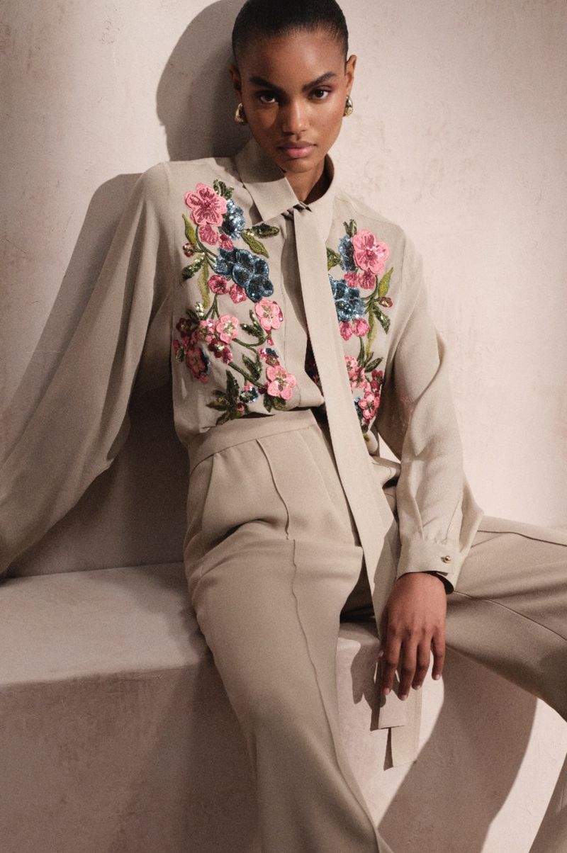 Elegant portrait of a woman in a button-down shirt and trousers, adorned with floral embellishments, against a neutral background.