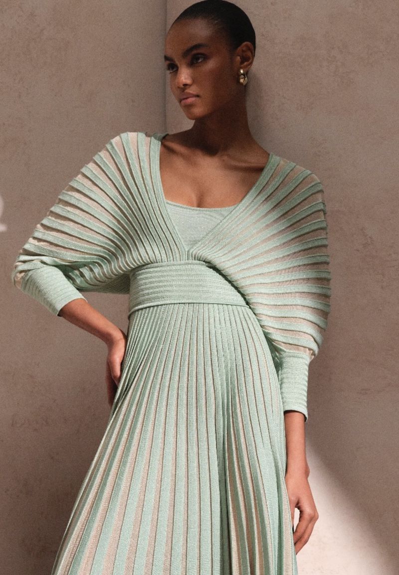 Elegant adult female with fair skin poses in a light green knit dress. Soft lighting and neutral background evoke a fashion editorial.