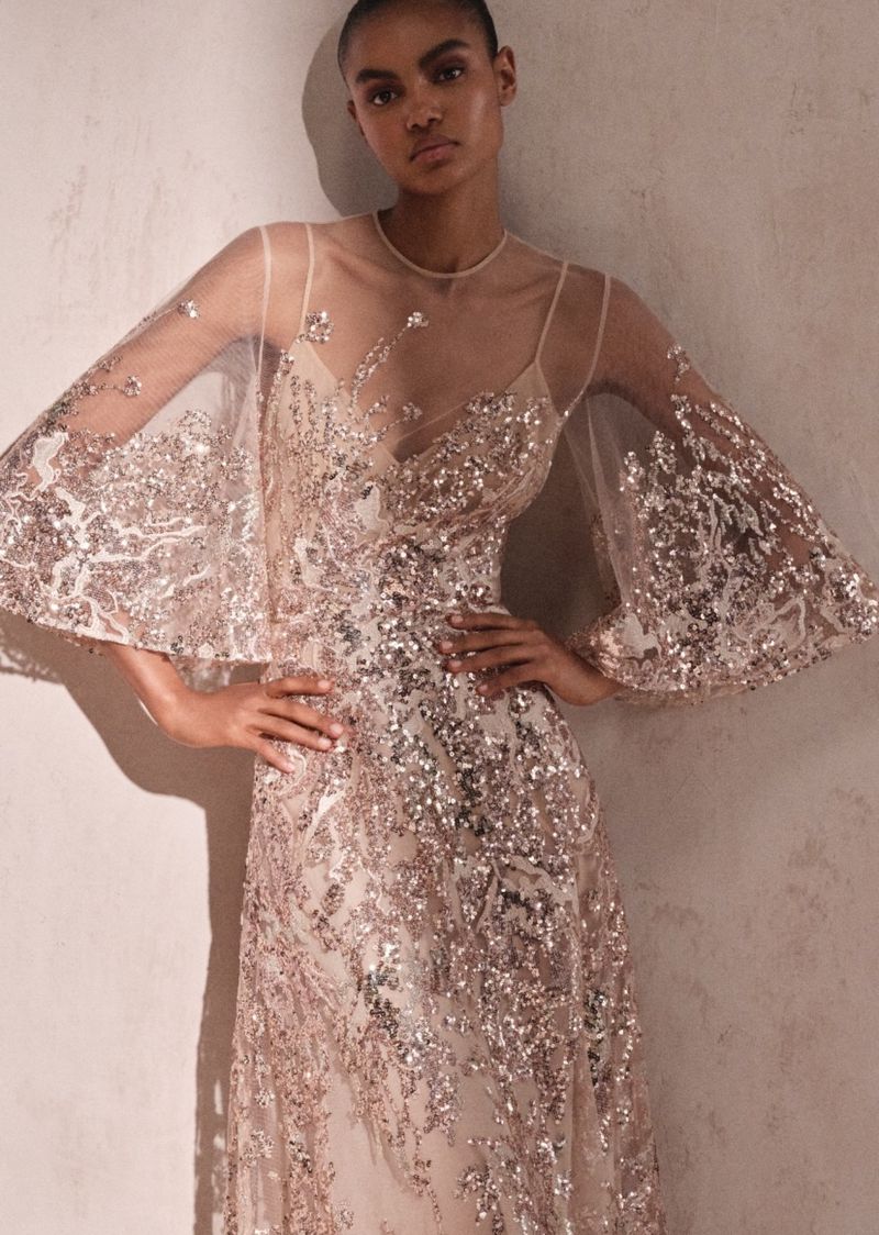 Elegant adult woman in a shimmering beaded and sequined formal gown with sheer sleeves, posing against a neutral-toned background.