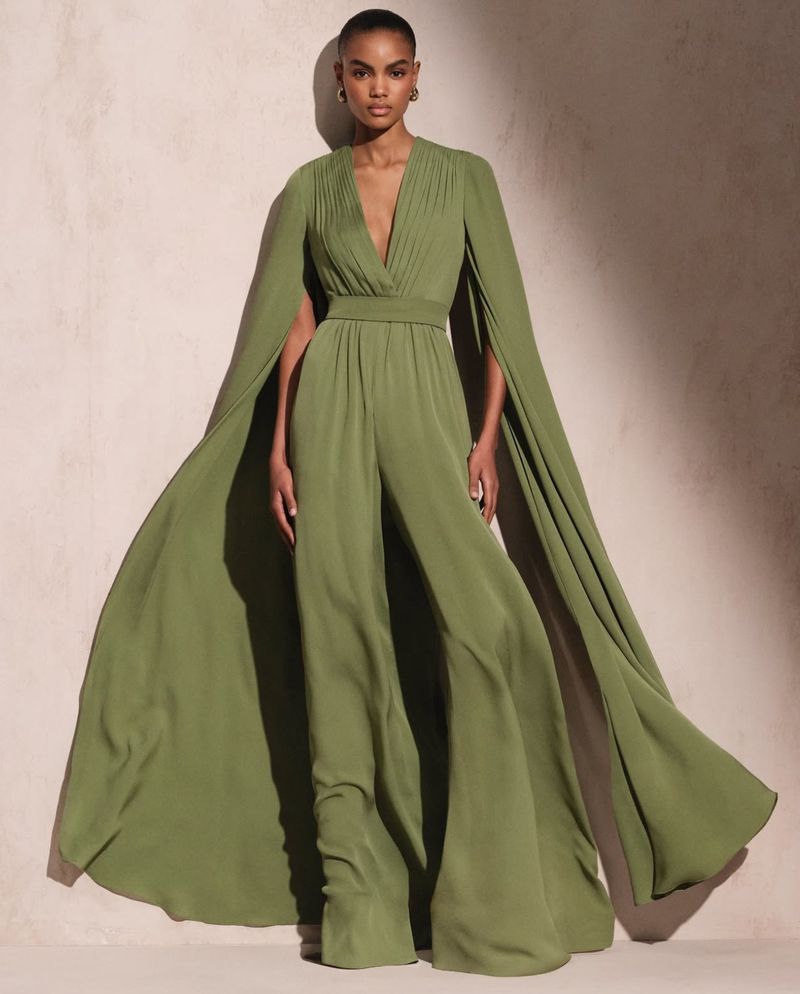 Elegant adult female poses in a flowing olive green jumpsuit with cape sleeves against a neutral backdrop in a studio setting.