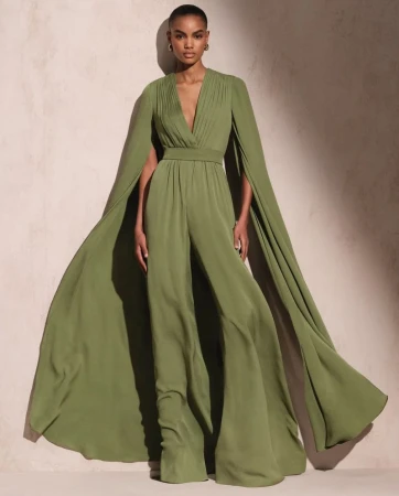 Elegant adult female poses in a flowing olive green jumpsuit with cape sleeves against a neutral backdrop in a studio setting.