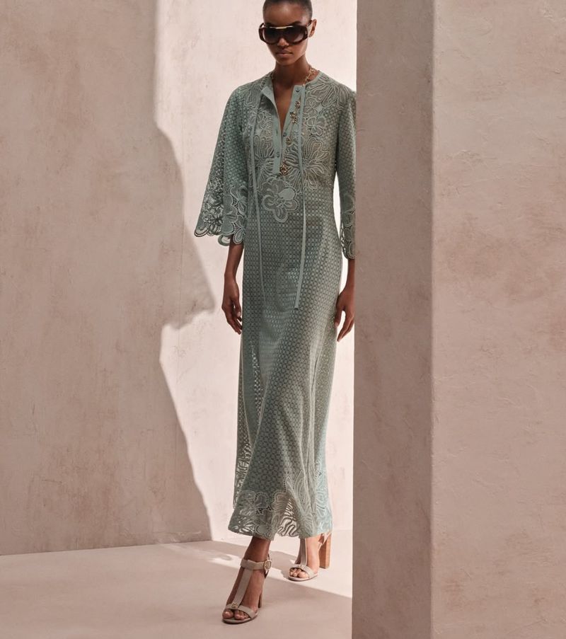 A woman poses in a full-length, sage-green, lace gown with semi-sheer sleeves, accessorized with sunglasses and heeled sandals.