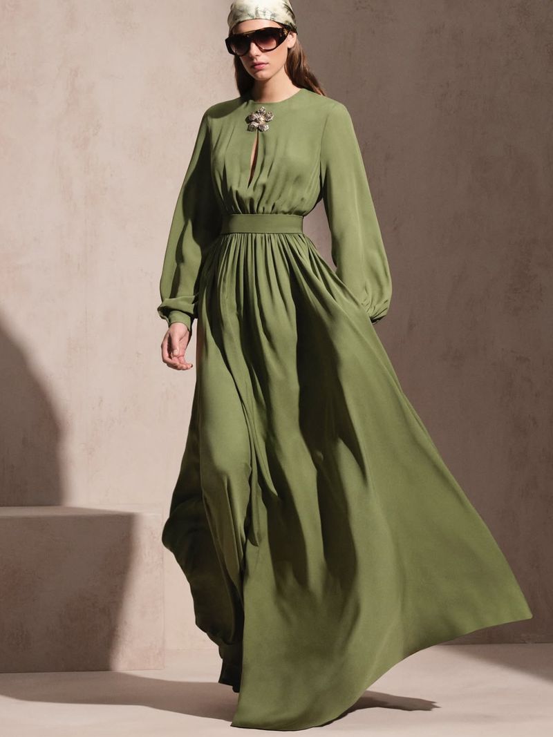 A woman stands in a studio with an olive-green gown, headscarf, and sunglasses in a full shot with a blurred background, fashion photography.
