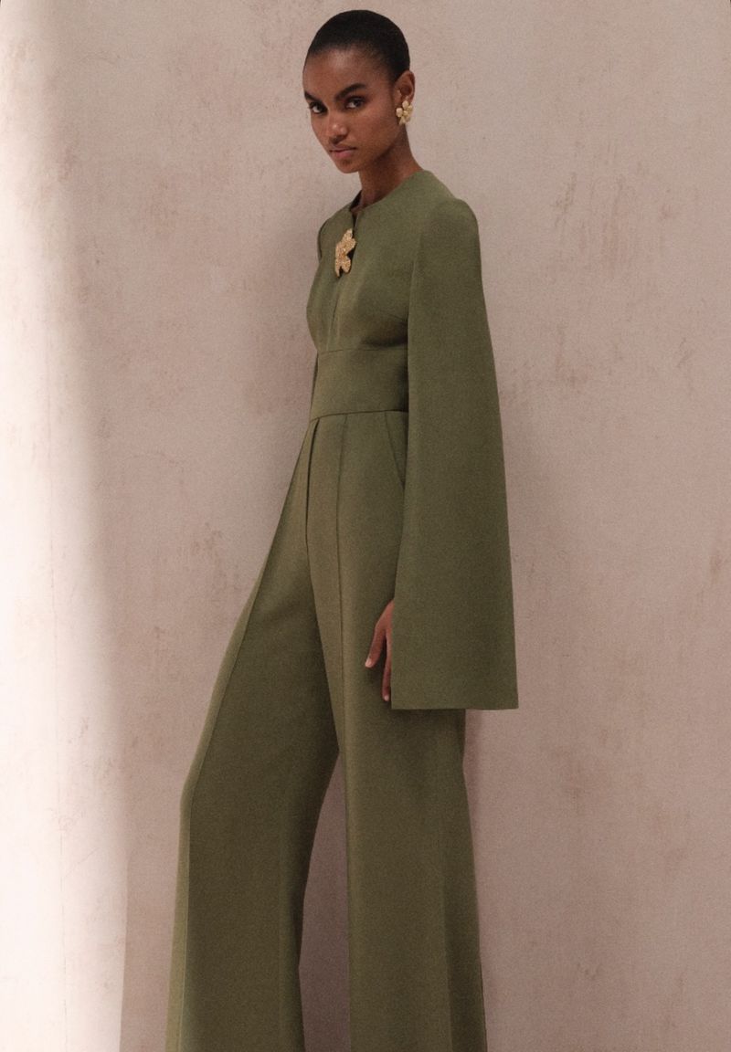 Woman in olive green jumpsuit with cape sleeves, posing in front of a neutral textured wall with soft, diffused light, medium shot.