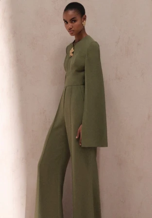 Woman in olive green jumpsuit with cape sleeves, posing in front of a neutral textured wall with soft, diffused light, medium shot.