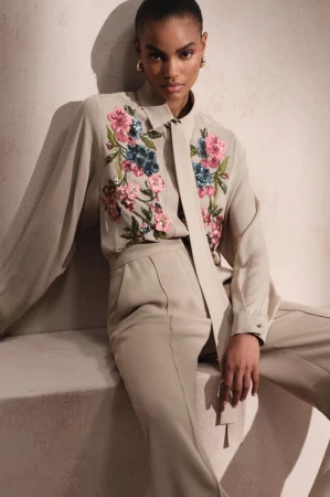 Model wearing an embroidered beige silk shirt and matching trousers seated against a neutral studio backdrop for Elie Saab.