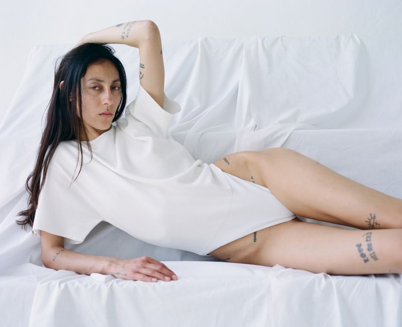 Woman lies on a white surface wearing a tee shirt with bare legs. The model has tattoos and dark hair and looks at the camera.