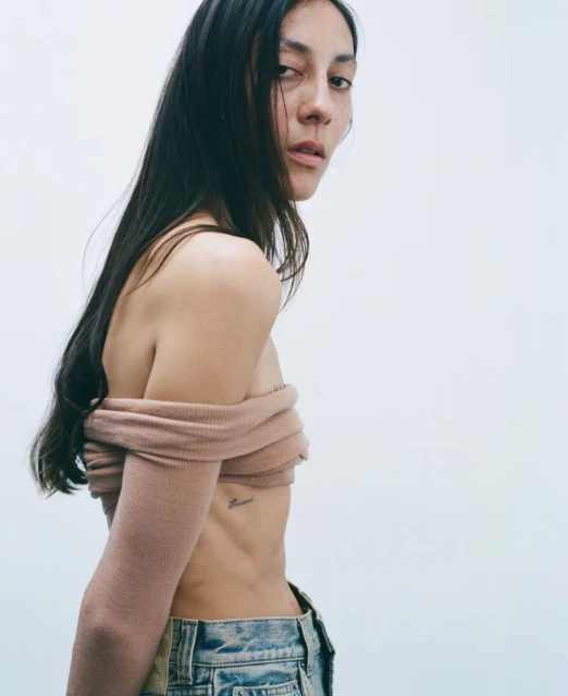 Attractive woman with long dark hair posing in blue jeans and a pink sweater against a bright white background in a studio portrait.