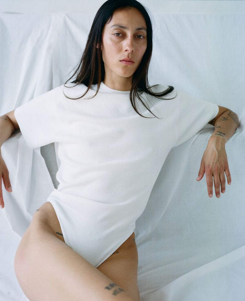 Model Gaia Orgeas wearing a white short-sleeve bodysuit reclining against draped white fabric backdrop.