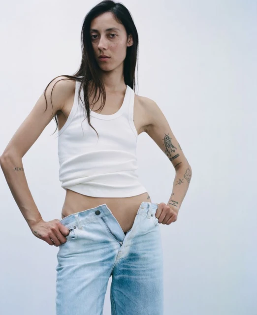 Portrait of a woman in white tank top and light blue jeans standing in a photo studio with a white background, fashion editorial.