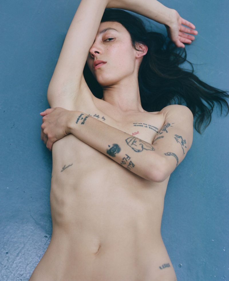 A slender person lies on a blue surface, gazing at the camera with a calm expression. The individual has multiple tattoos on their arms and torso.