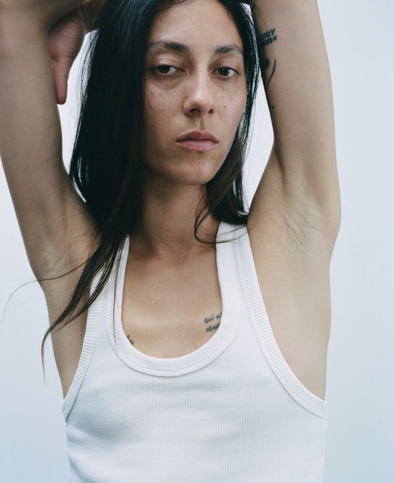 Close-up of a brunette woman in a white tank top with her arms raised. She poses against a plain light-colored background.