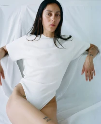 Model Gaia Orgeas wearing a white short-sleeve bodysuit reclining against draped white fabric backdrop.