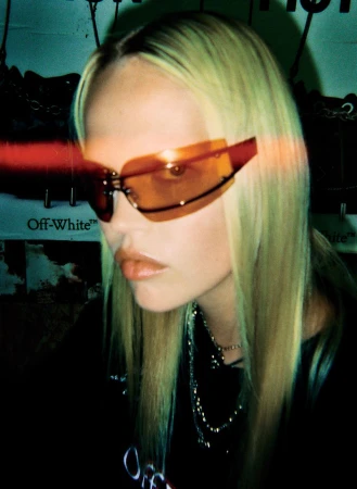Blonde woman wearing orange futuristic sunglasses and silver necklaces poses indoors against a backdrop with fashion brand logos.