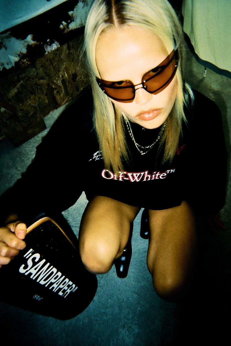 High-angle shot of a blonde woman with sunglasses crouching and holding a black can with 'SANDPAPER' printed on the side indoors.
