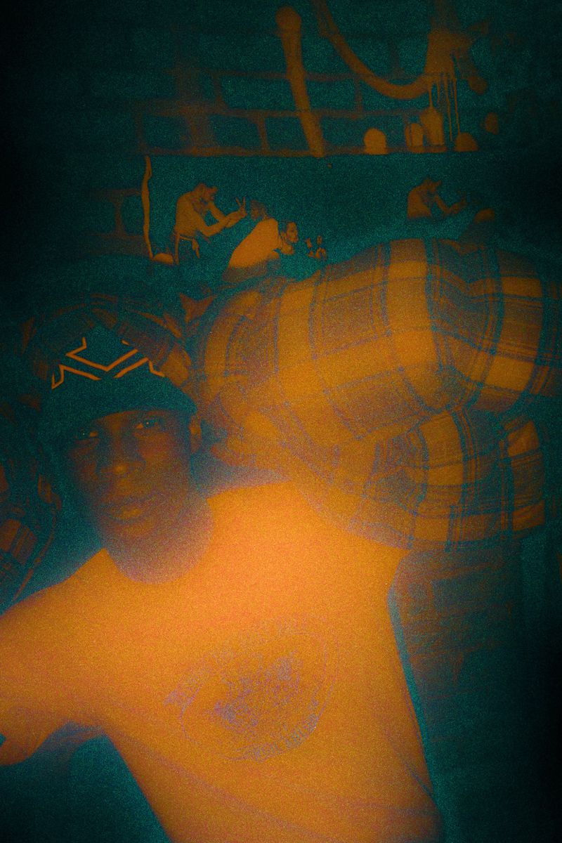 Portrait of an adult in a t-shirt and bandana, standing indoors with blurred background and moody lighting with a grainy texture.