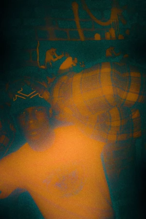Portrait of an adult in a t-shirt and bandana, standing indoors with blurred background and moody lighting with a grainy texture.