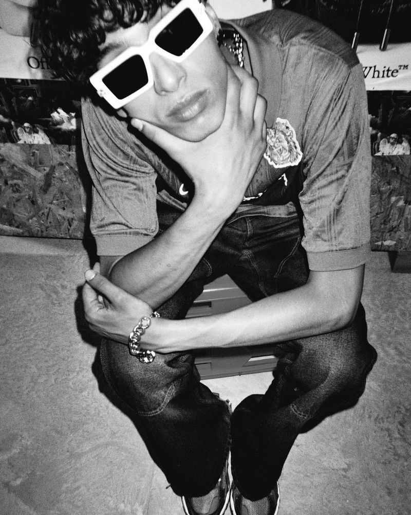 Black and white portrait of a young adult male seated with sunglasses, stylish attire, and casual pose, indoors with natural lighting.