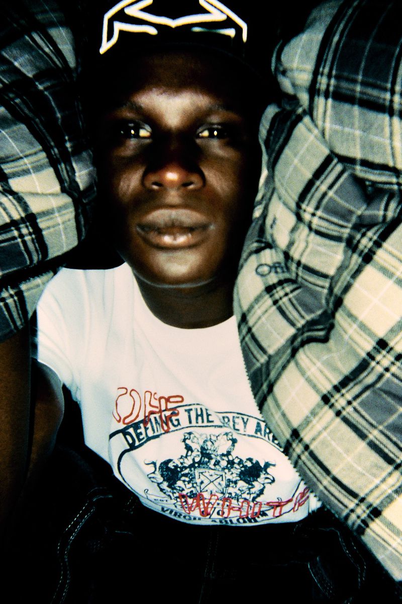 Portrait of a dark-skinned adult in a white tee shirt with graphic text, partially framed by a plaid pillow and a glowing crown shape.