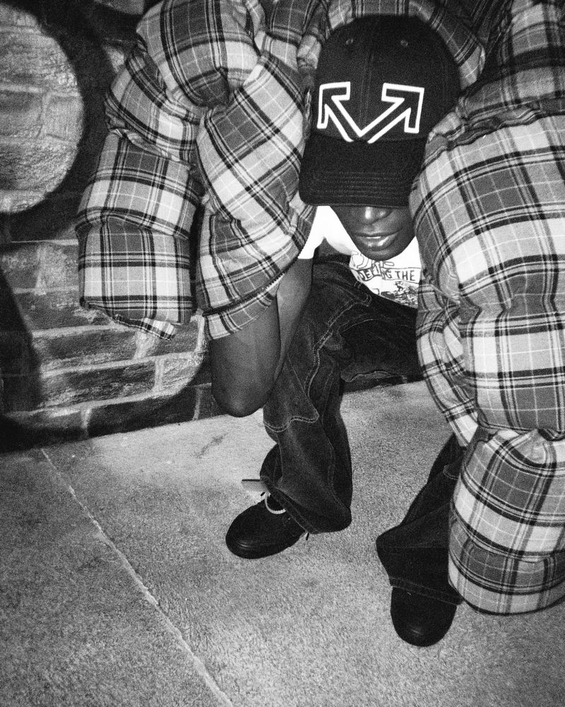 A young adult crouches in a plaid shirt jacket and cap in a grayscale image, creating an urban, casual fashion shot.
