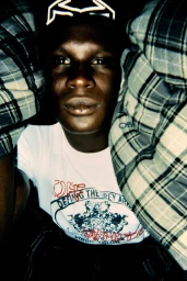 Close-up portrait of a Black man wearing a white graphic t-shirt framed by green and grey plaid fabric.