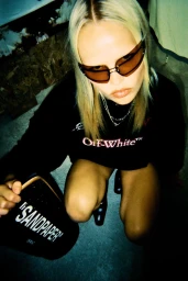 Blonde woman in brown wrap sunglasses crouching in a black Off-White sweatshirt holding a black Sandpaper bag.