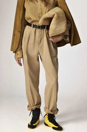 Fashionable adult woman posing in a studio setting, wearing brown pants and jacket with yellow and black sneakers.