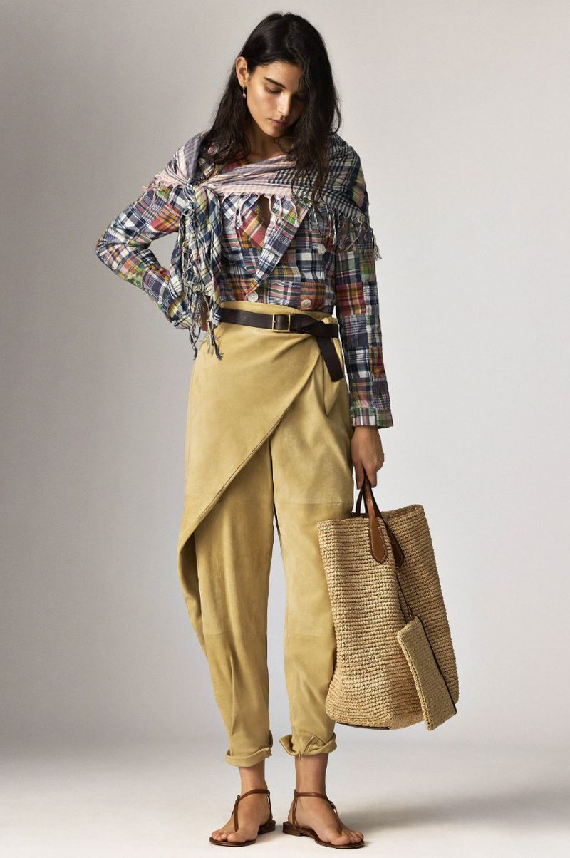 A woman models a colorful plaid top, tan wrap pants, flat sandals, and a woven straw bag in a studio setting for a fashion editorial.