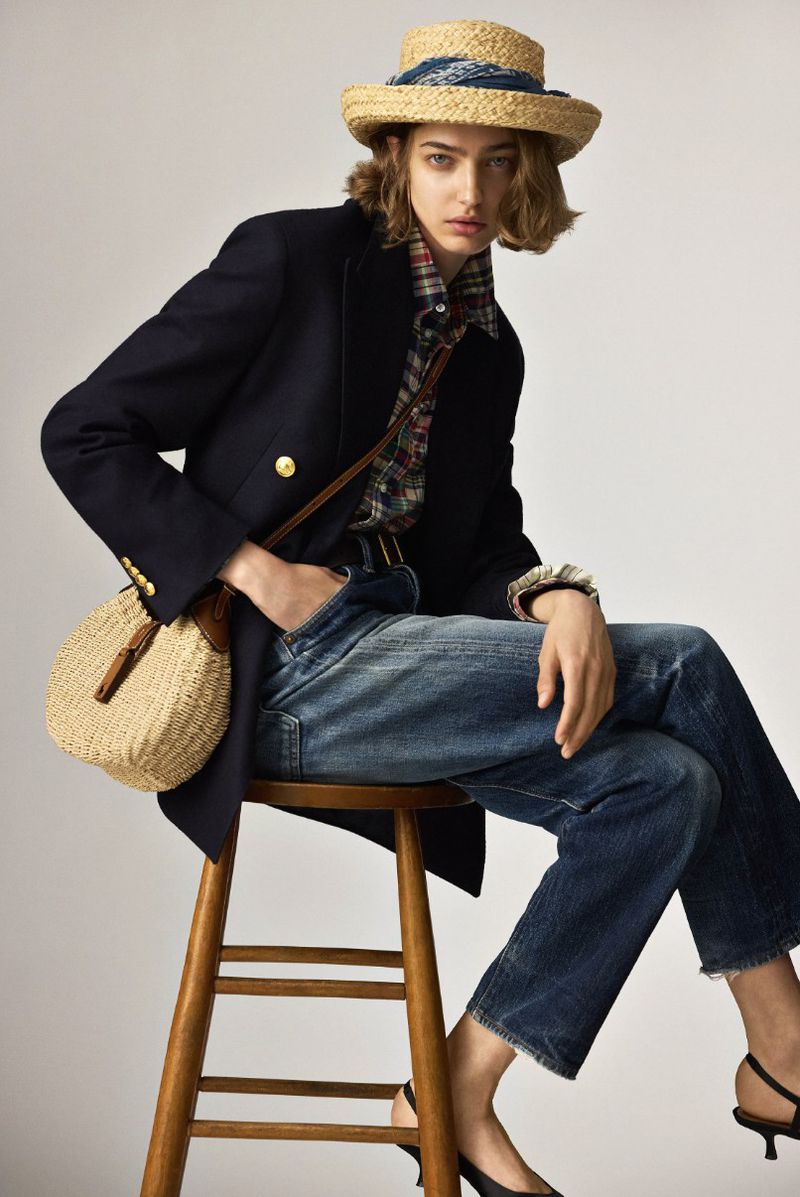 Fashionable woman sits on a stool, wearing a straw hat, navy blazer, plaid shirt, and jeans, paired with black heels and a straw bag.