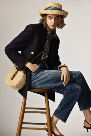 Fashionable woman sits on a stool, wearing a straw hat, navy blazer, plaid shirt, and jeans, paired with black heels and a straw bag.