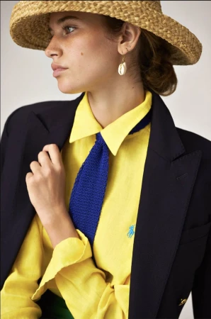 Woman in yellow shirt, blue tie, and black blazer wearing straw hat and cowrie shell earring. Indoor studio portrait fashion shot.