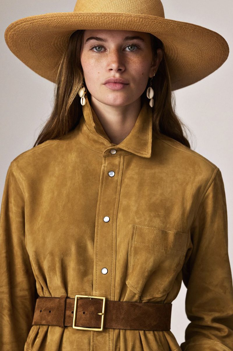 Portrait of a woman wearing a straw hat, collared shirt, and belt with shell earrings, posing in a light studio setting.