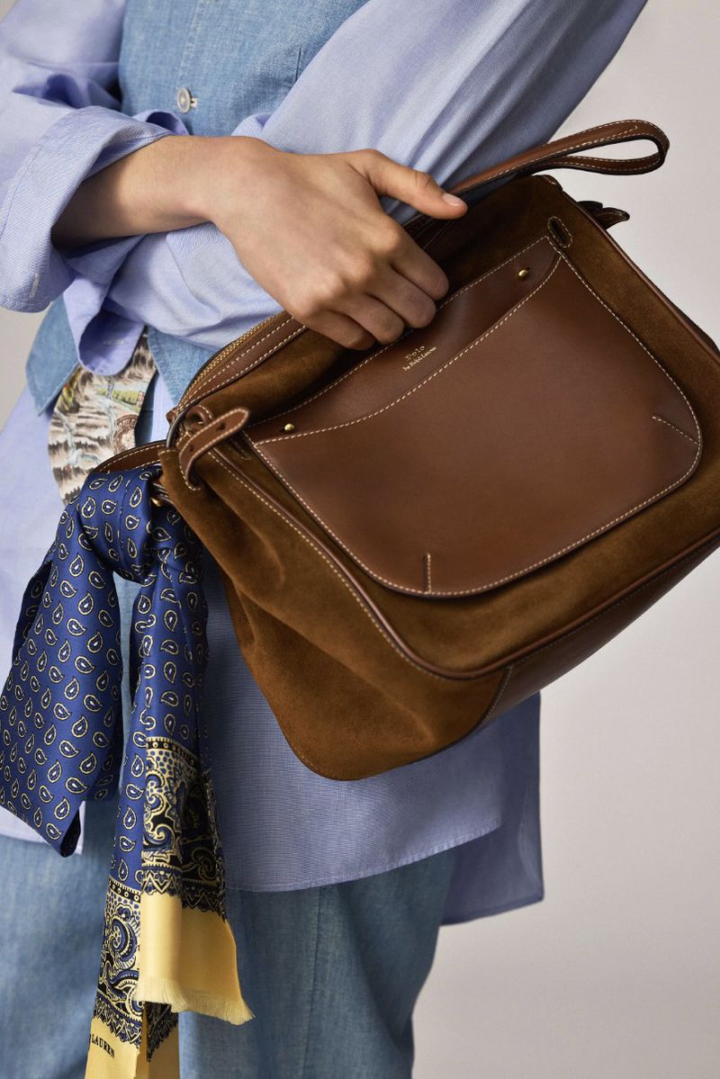 Stylish brown leather shoulder bag held by a person wearing a blue blouse and jeans, accessorized with a patterned blue and yellow scarf.