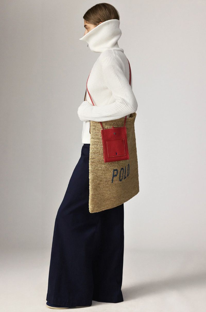 A woman in a white turtleneck sweater and navy pants poses with a straw tote bag featuring a red pocket and Polo branding.