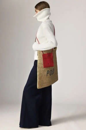 A woman in a white turtleneck sweater and navy pants poses with a straw tote bag featuring a red pocket and Polo branding.