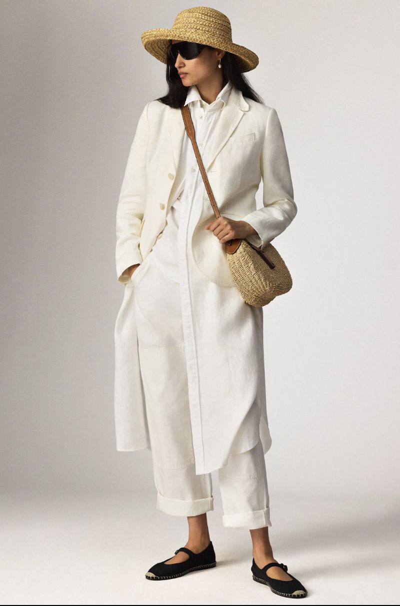 Elegant woman in a white linen coat, shirt, and pants, accessorized with a straw hat, woven bag, and black flats against a neutral backdrop.