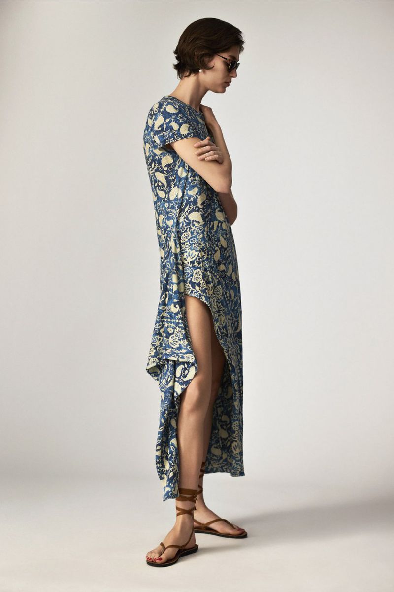 A woman models a blue and beige floral maxi dress with a thigh-high slit, paired with strappy brown sandals, in a studio setting.