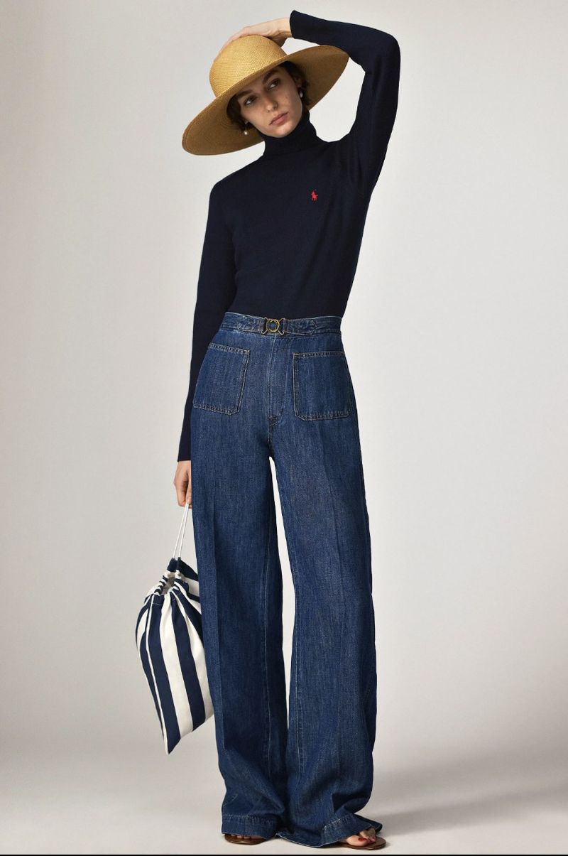 Fashion shot of a woman wearing a navy turtleneck, wide-leg jeans, straw hat, and carrying a striped bag in a studio setting.