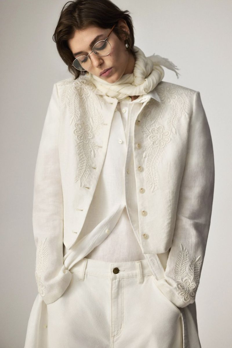 Adult woman with short brown hair, wearing white jacket and pants, round glasses, and white scarf, posing in a light studio setting.