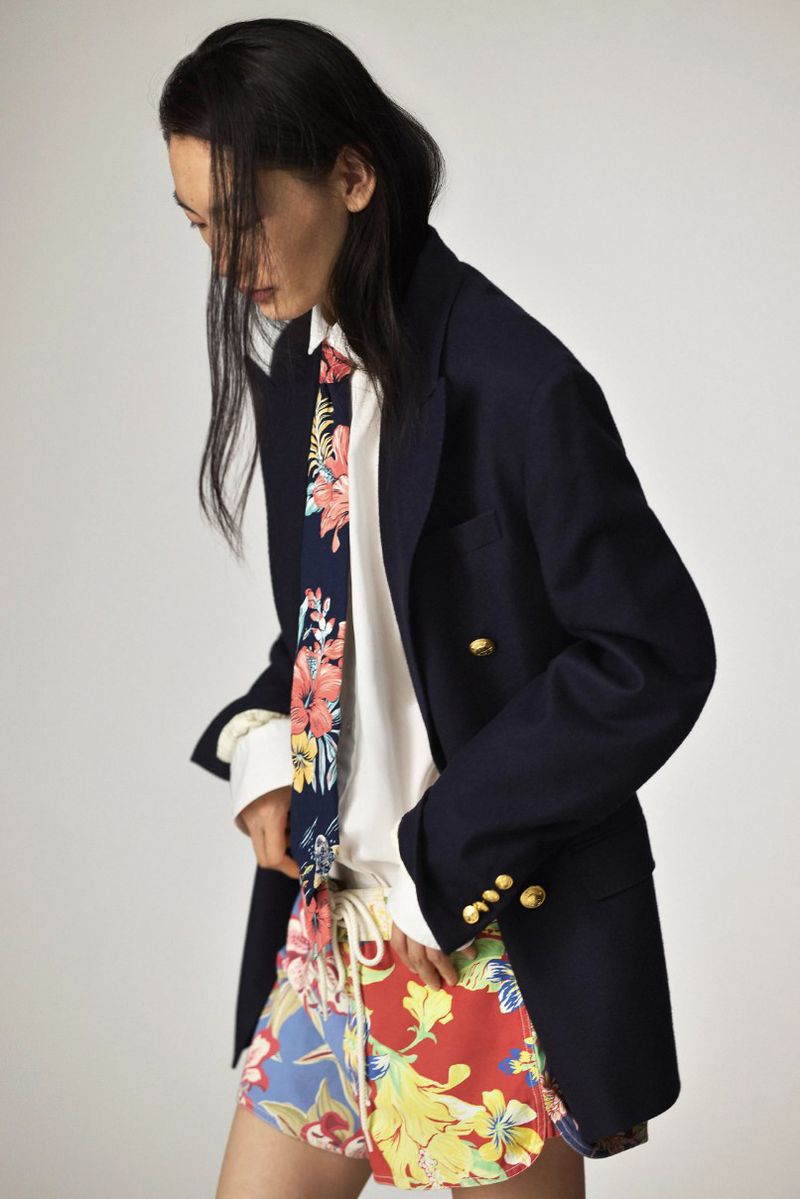 Portrait of a slender person in an oversized navy blazer, white button-down, floral tie, and floral shorts against a neutral backdrop.