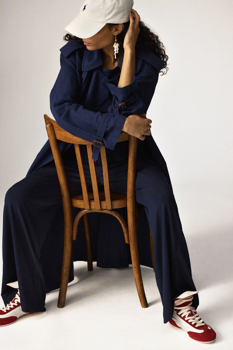 A dark-haired woman sits on a wooden chair in a photo studio wearing an oversized blue trench coat, sweatpants, red sneakers, and a white baseball cap.