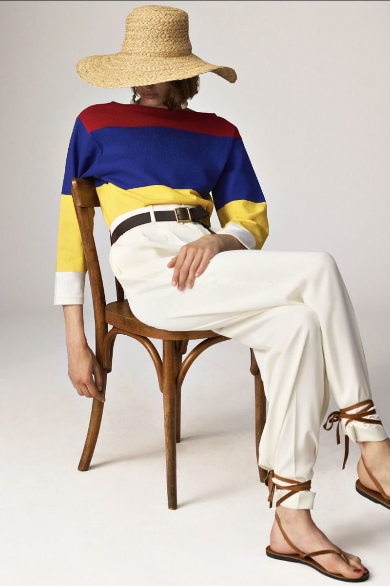 Woman poses in color-blocked shirt, white pants, sandals, and straw hat while seated on wood chair against a neutral backdrop.