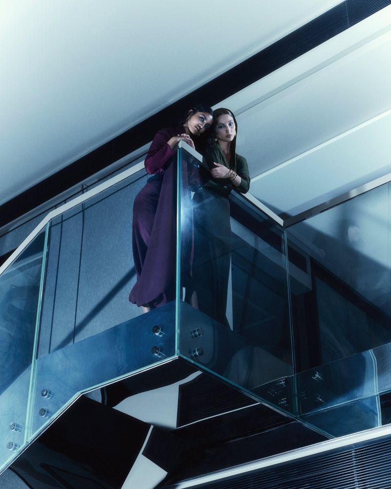 Two women stand on a glass staircase in an indoor setting, wearing fashionable clothing, captured from a low angle perspective with blue tones.