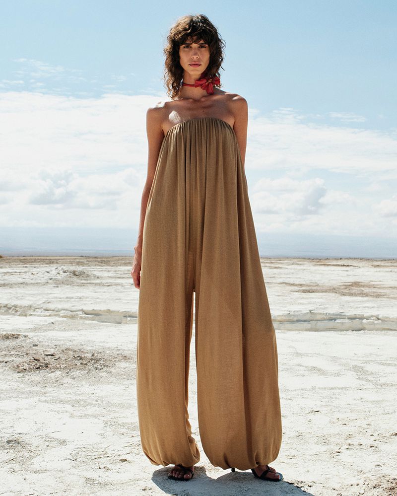 A woman stands in a neutral-toned jumpsuit with a red scarf, posing in a sunlit, arid landscape under a blue sky with clouds.