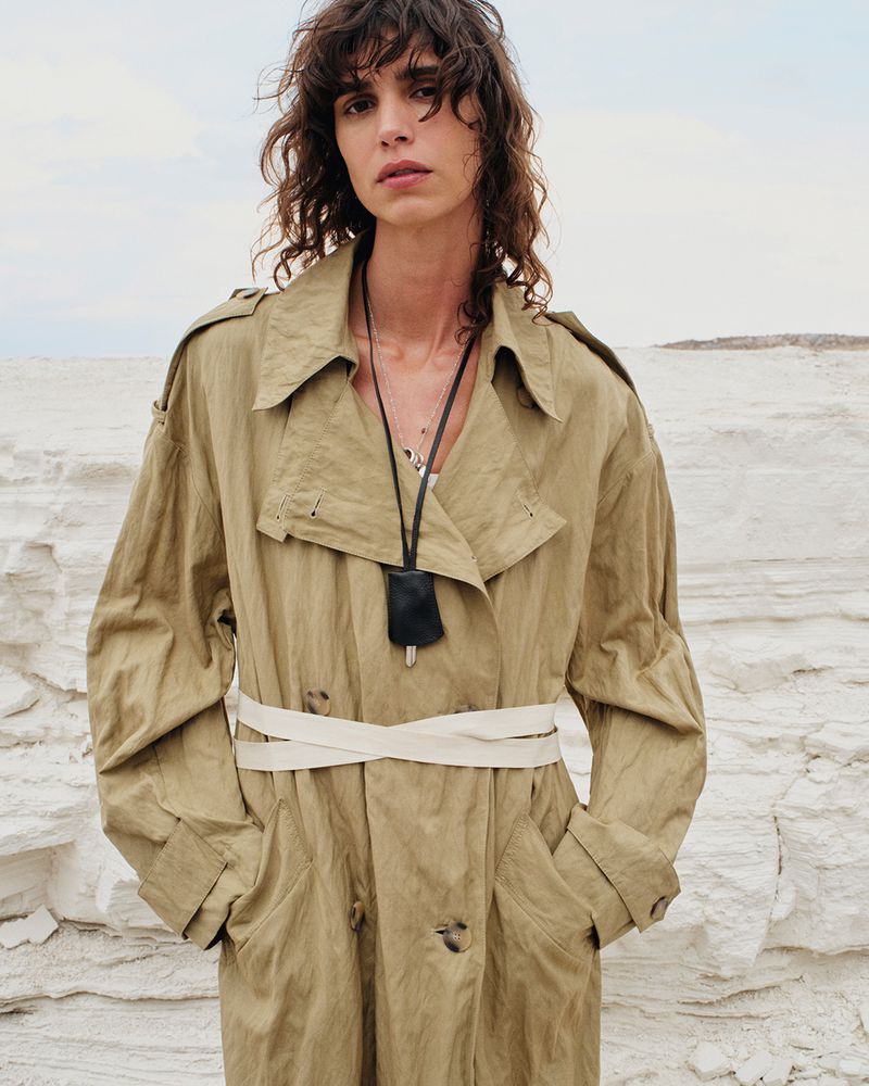 Woman in a khaki trench coat with a white belt, standing in a rocky environment with a cloudy sky in the background.