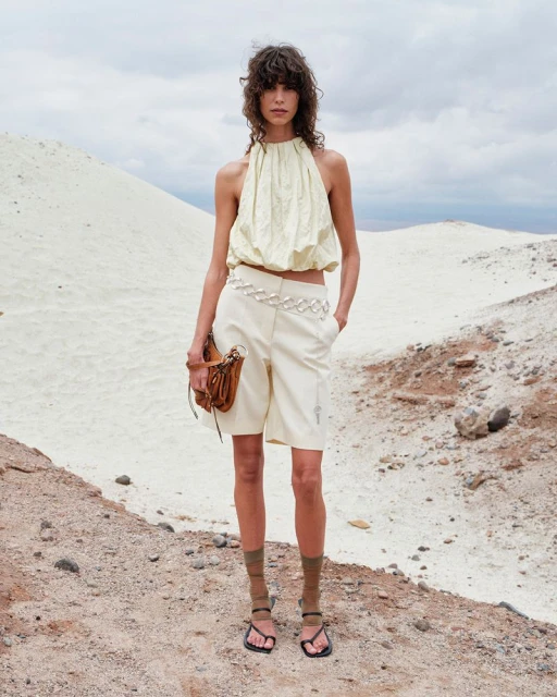 Woman in a cream-colored halter top and shorts poses in an arid landscape with brown purse, creating a fashion editorial image.