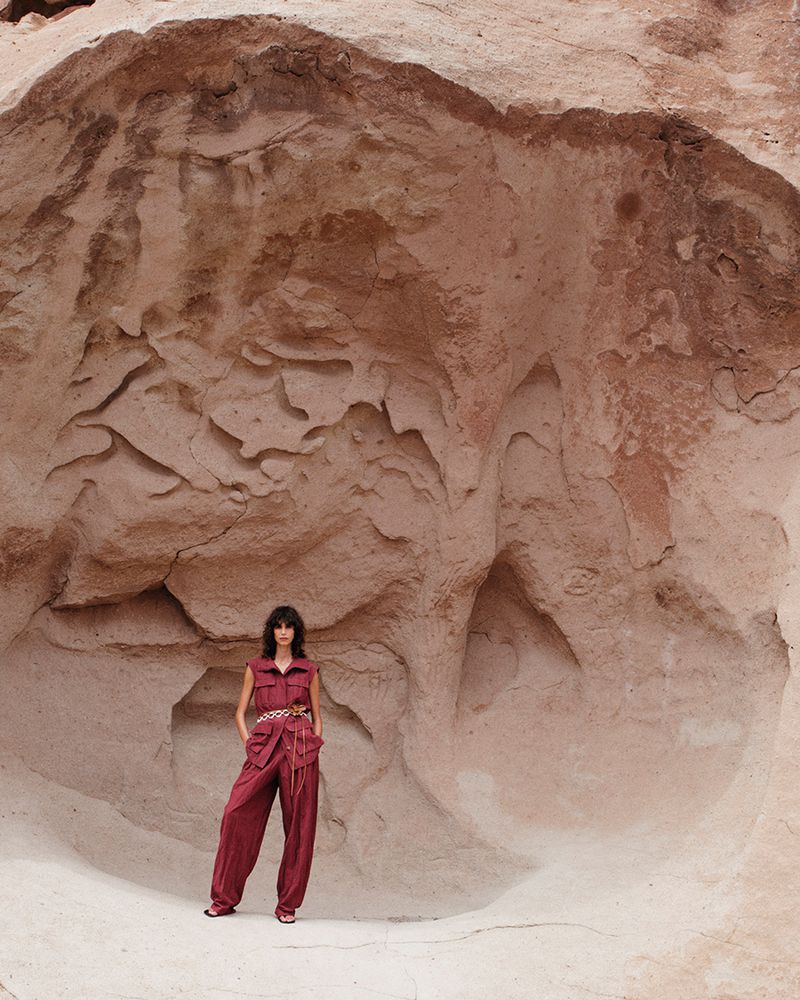 Woman in a red jumpsuit poses in a cave with textured sandstone walls, wearing a tied belt and sandals for a fashion shot.