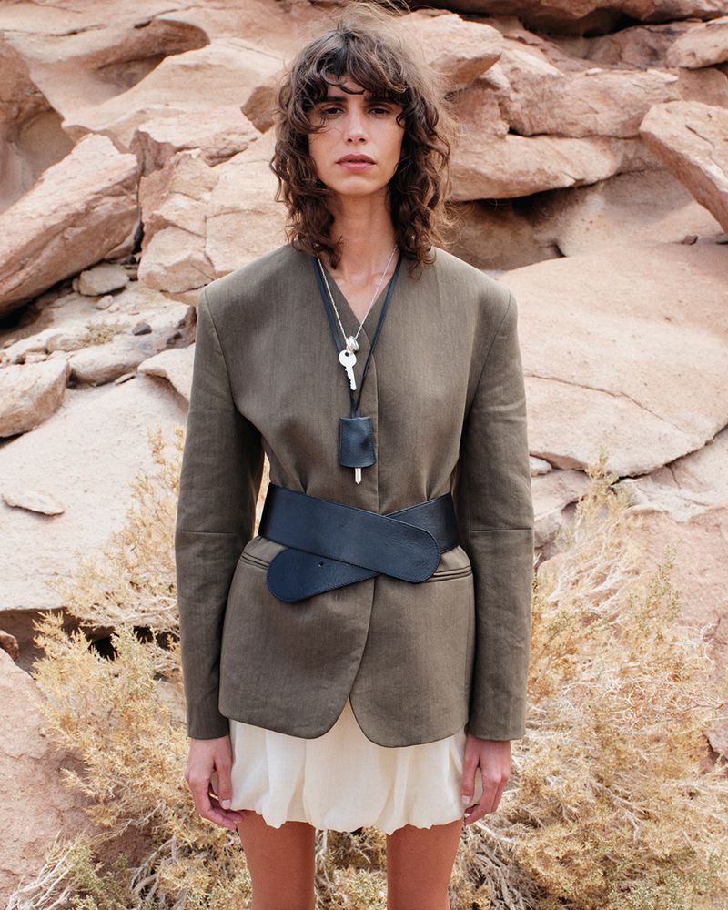 An adult woman poses in a brown blazer, black belt, and light skirt, outdoors amid desert vegetation and rock formations on a sunny day.