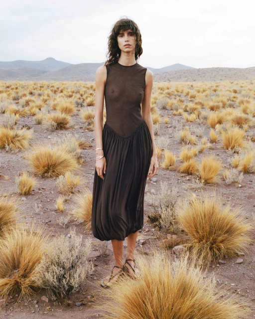Adult female fashion model in a dark dress stands in a desert-like landscape with low shrubs and distant mountains under an overcast sky.
